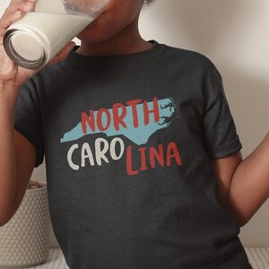 May include: A young person wearing a dark blue t-shirt with a white outline of North Carolina and the text "NORTH CAROLINA" in red and blue. They are drinking from a glass of milk and holding a chocolate chip cookie.