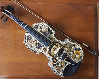 May include: A black violin decorated with white pearls, gold beads, and other embellishments. The violin is lying on a wooden surface with its bow beside it.