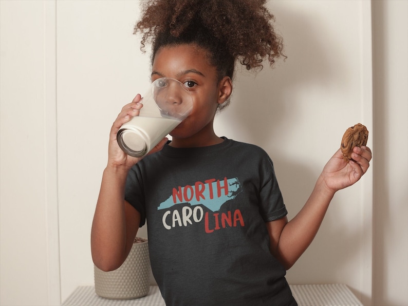 May include: A young person wearing a dark blue t-shirt with a white outline of North Carolina and the text "NORTH CAROLINA" in red and blue. They are drinking from a glass of milk and holding a chocolate chip cookie.