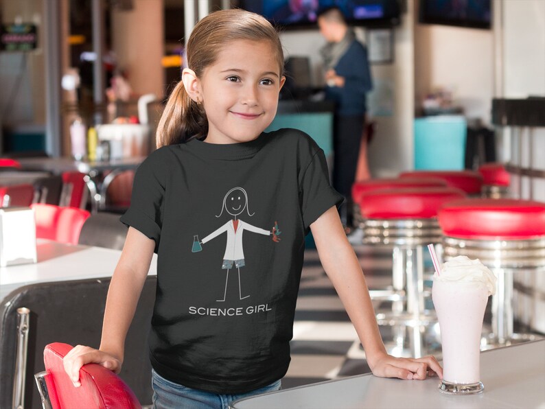 Puede incluir: Una persona joven lleva una camiseta negra con una ilustraci&oacute;n de un cient&iacute;fico de palo blanco sosteniendo un vaso de precipitados y un tubo de ensayo. La camiseta tiene el texto "SCIENCE GIRL" impreso en la parte inferior.