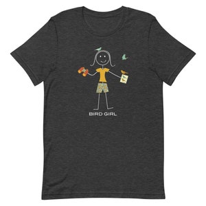 May include: A dark heather grey t-shirt with a stick figure illustration of a person wearing a yellow shirt, patterned shorts, and holding binoculars and a field guide. The text "BIRD GIRL" is printed below the figure.