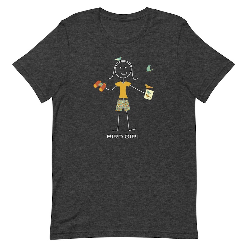 May include: A dark heather grey t-shirt with a stick figure illustration of a person wearing a yellow shirt, patterned shorts, and holding binoculars and a field guide. The text "BIRD GIRL" is printed below the figure.
