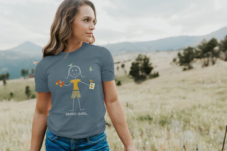 May include: A woman wearing a blue t-shirt with a stick figure illustration of a person holding a bird feeder and a book. The text "BIRD GIRL" is printed below the illustration.