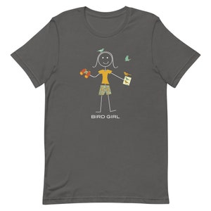 May include: A dark gray t-shirt with a stick figure illustration of a person holding binoculars and a bird guide. The figure is surrounded by small birds. The text "BIRD GIRL" is printed below the figure.