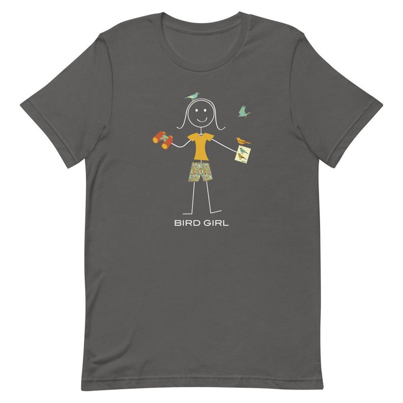 May include: A dark gray t-shirt with a stick figure illustration of a person holding binoculars and a bird guide. The figure is surrounded by small birds. The text "BIRD GIRL" is printed below the figure.