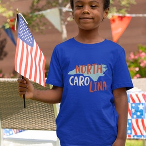 May include: A young person wearing a blue t-shirt with the text "NORTH CAROLINA" and an outline of the state in blue. The person is holding a small American flag.