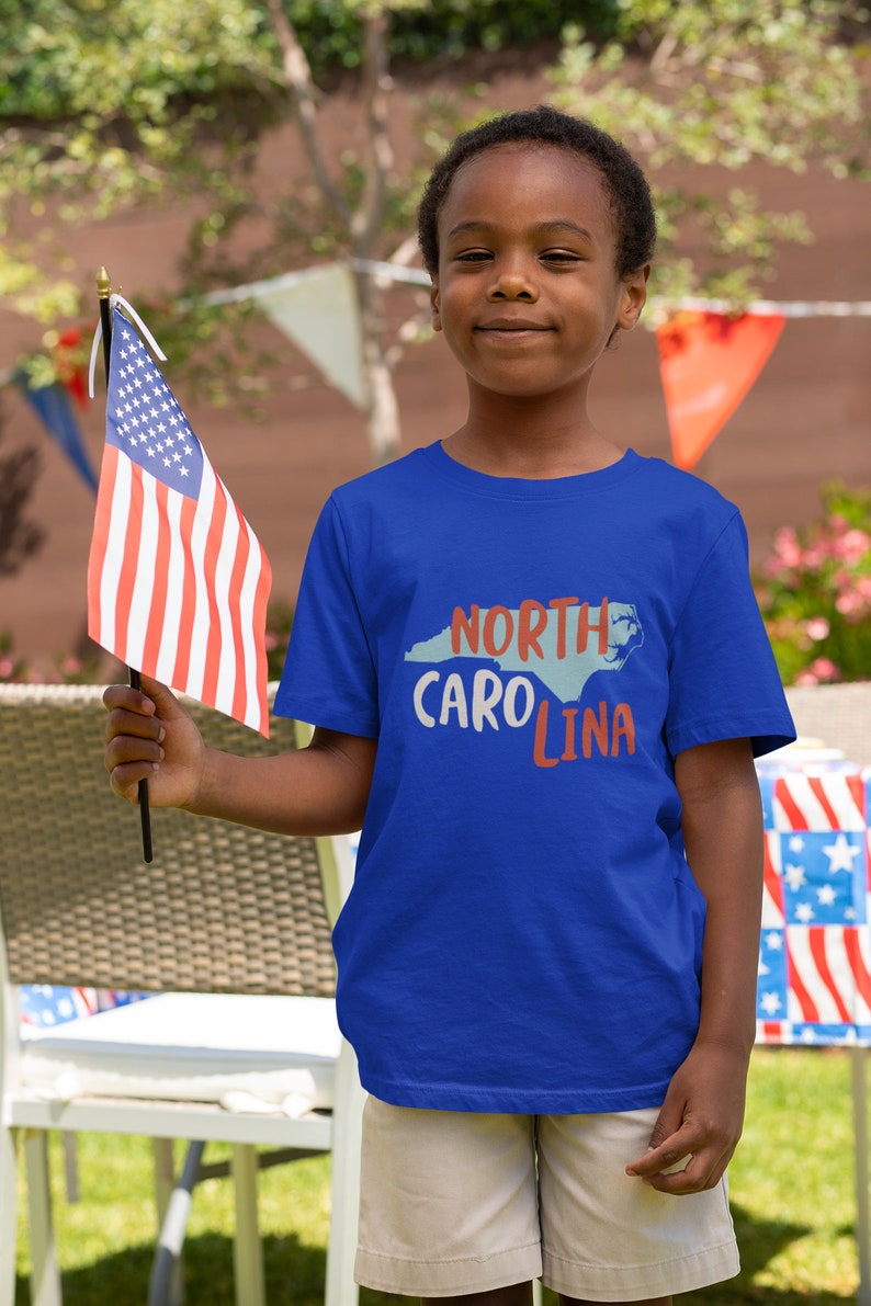 May include: A young person wearing a blue t-shirt with the text "NORTH CAROLINA" and an outline of the state in blue. The person is holding a small American flag.