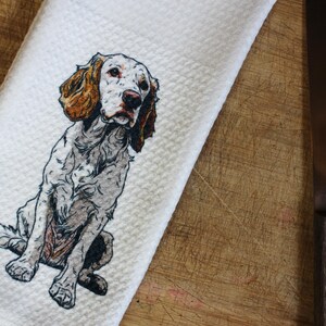 May include: White kitchen towel featuring a detailed illustration of a seated English Setter dog. The dog has brown and white markings. The towel is on a wooden cutting board with a stainless steel knife.