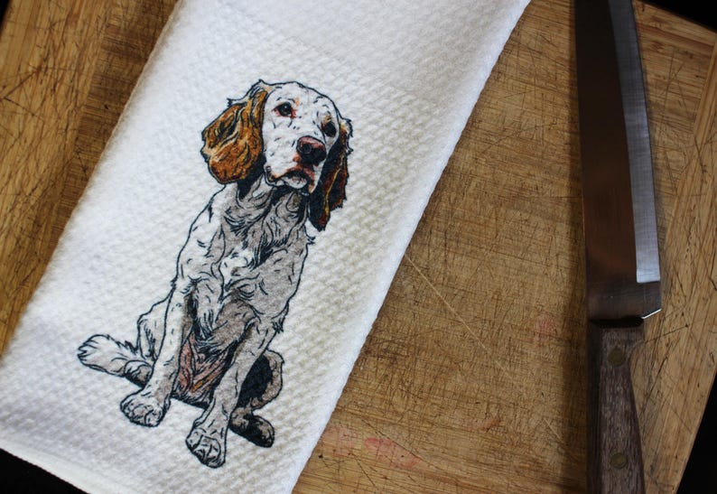 May include: White kitchen towel featuring a detailed illustration of a seated English Setter dog. The dog has brown and white markings. The towel is on a wooden cutting board with a stainless steel knife.