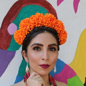 Puede incluir: Una mujer con una blusa blanca de hombros descubiertos con encaje, un collar de cuentas de colores y una corona de flores. Está de pie frente a una pared colorida.
