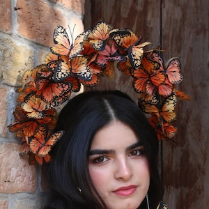 Puede incluir: Un tocado estilo halo con numerosas réplicas de mariposas monarca naranjas y negras, combinado con pendientes de mariposa a juego y un collar de varias hileras compuesto por cuentas redondas moteadas en azul y marrón.