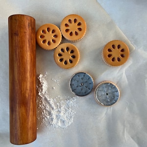 Puede incluir: Primer plano de un rodillo de madera y varias galletas redondas con glaseado de azúcar moreno. Las galletas están dispuestas sobre una superficie blanca con un espolvoreo de azúcar en polvo.