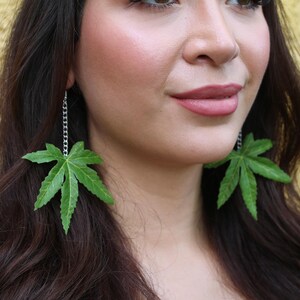 May include: Green leaf earrings. Each earring features a large, detailed green leaf pendant suspended from a silver chain. The leaves have a naturalistic appearance with intricate veining, creating a unique and eye-catching accessory.