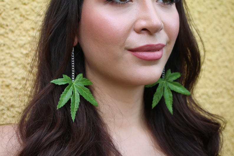 May include: Green leaf earrings. Each earring features a large, detailed green leaf pendant suspended from a silver chain. The leaves have a naturalistic appearance with intricate veining, creating a unique and eye-catching accessory.