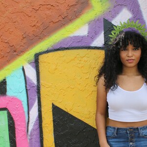 May include: A person wearing a white spaghetti strap crop top and blue denim jeans stands in front of a colorful graffiti wall. A green leaf crown adorns the person's curly black hair.