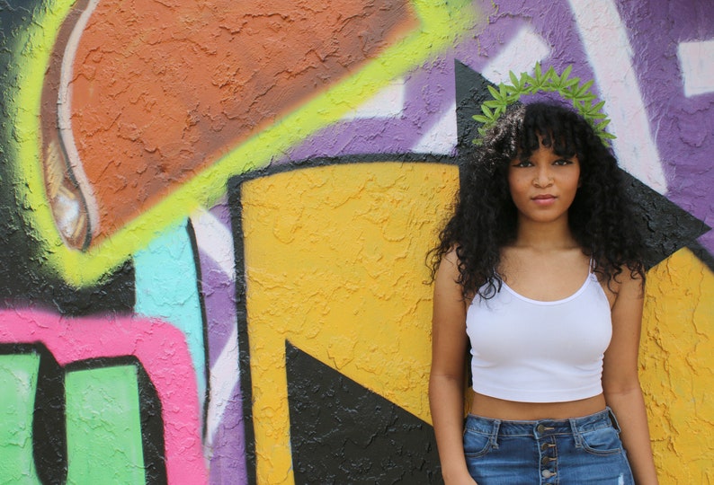 May include: A person wearing a white spaghetti strap crop top and blue denim jeans stands in front of a colorful graffiti wall. A green leaf crown adorns the person's curly black hair.