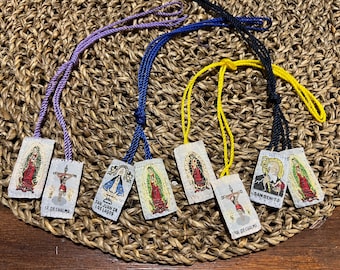 May include: Seven colorful religious medallions with cords, each featuring a different saint or religious figure. The medallions are on a woven straw background. The medallions feature images of Saint Juan de los Lagos, Saint Benito, Our Lady of Chalma, and others.