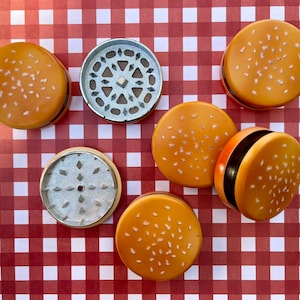 May include: Six metal grinders shaped like hamburger buns with sesame seeds. The grinders are silver and have a textured surface.