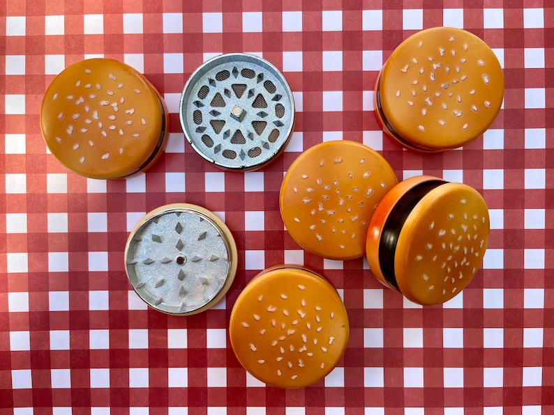 May include: Six metal grinders shaped like hamburger buns with sesame seeds. The grinders are silver and have a textured surface.