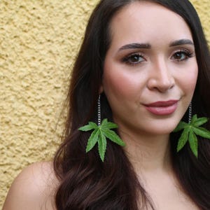 May include: Green leaf earrings with a silver chain dangle. The earrings are a vibrant green color and are shaped like cannabis leaves. The woman in the image has long dark hair and is wearing makeup.