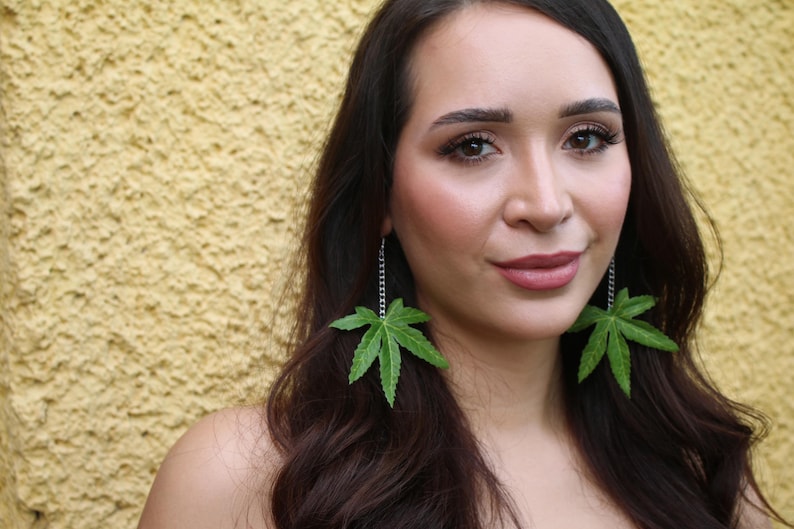 May include: Green leaf earrings with a silver chain dangle. The earrings are a vibrant green color and are shaped like cannabis leaves. The woman in the image has long dark hair and is wearing makeup.