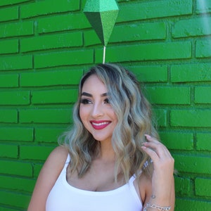 Puede incluir: Una mujer con el pelo largo y rubio lleva un top blanco de tirantes y una diadema verde en forma de diamante. Est&aacute; sonriendo y mirando hacia un lado. El fondo es una pared de ladrillo verde.