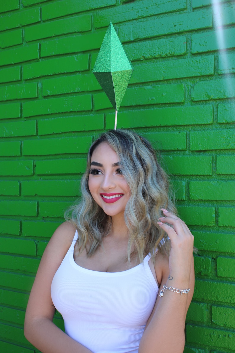 Puede incluir: Una mujer con el pelo largo y rubio lleva un top blanco de tirantes y una diadema verde en forma de diamante. Est&aacute; sonriendo y mirando hacia un lado. El fondo es una pared de ladrillo verde.