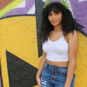 May include: A woman wearing a white crop top and blue jeans with distressed patches. She is wearing a green leaf crown. The background is a colorful graffiti wall with yellow, black, and purple paint.