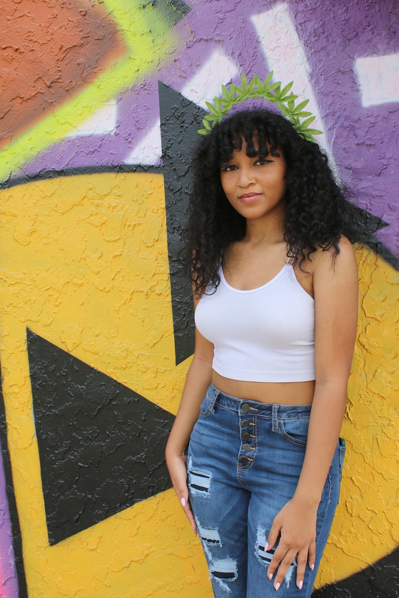 May include: A woman wearing a white crop top and blue jeans with distressed patches. She is wearing a green leaf crown. The background is a colorful graffiti wall with yellow, black, and purple paint.