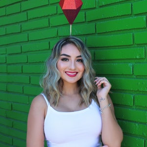 Puede incluir: Una mujer con cabello rubio que lleva un top blanco sin mangas y jeans azules. Est&aacute; sonriendo y tiene un objeto rojo en forma de diamante en la cabeza.