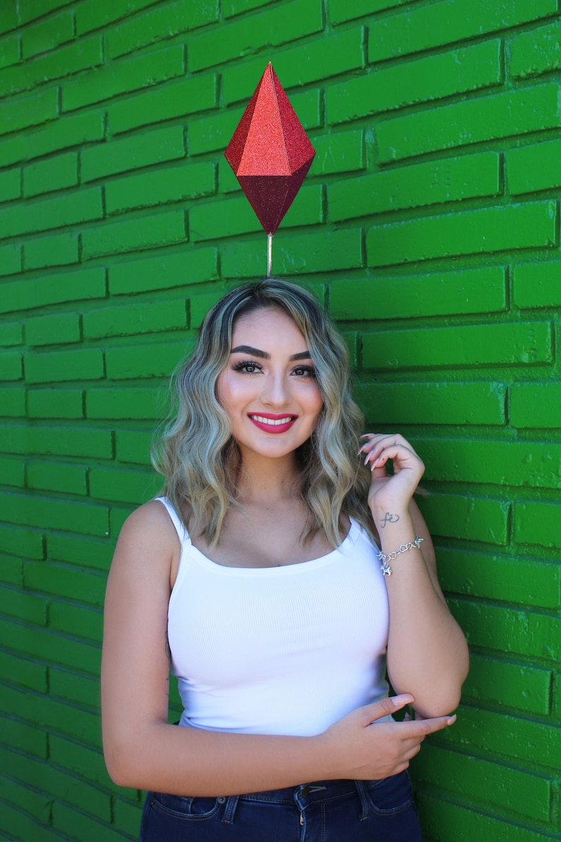 Puede incluir: Una mujer con cabello rubio que lleva un top blanco sin mangas y jeans azules. Est&aacute; sonriendo y tiene un objeto rojo en forma de diamante en la cabeza.