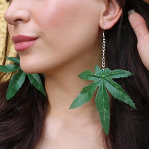 May include: Green leaf-shaped earrings. The earrings are made of a dark green material and hang from silver-colored chains. The leaves have detailed veining and are a vibrant green color.