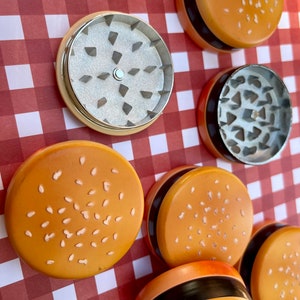 May include: A set of six grinder pieces shaped like hamburger buns with sesame seeds. The grinders are made of metal and have a black ring around the top. The grinders are on a red and white checked background.