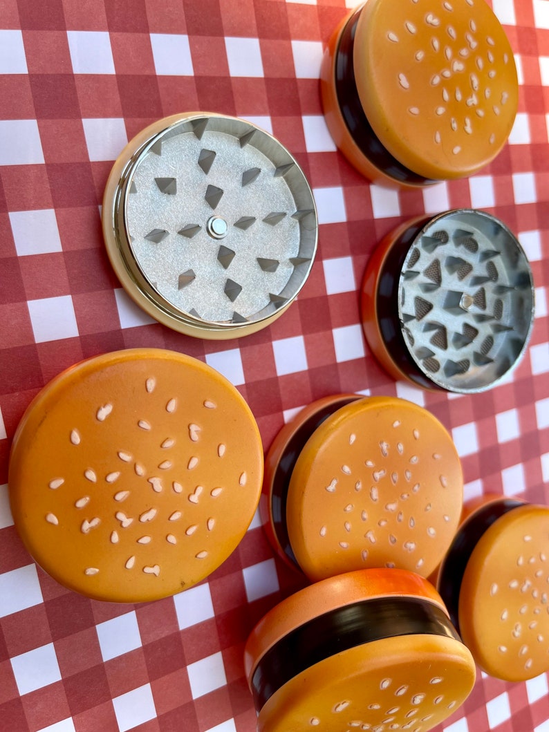 May include: A set of six grinder pieces shaped like hamburger buns with sesame seeds. The grinders are made of metal and have a black ring around the top. The grinders are on a red and white checked background.