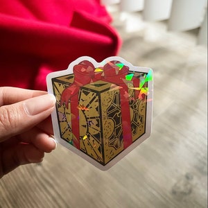 May include: A holographic sticker of a gold gift box with a red ribbon. The box is decorated with intricate details and has a holographic finish.