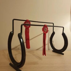 May include: A black metal horseshoe-shaped rack with two red floral accents and tassels. The rack has a horizontal bar for hanging items. The design incorporates two upright horseshoe shapes and a third horseshoe shape on the right side.