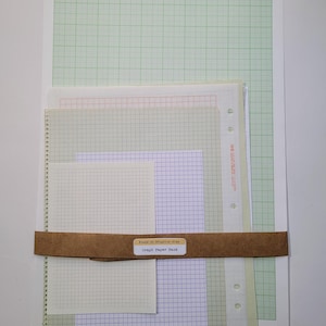 May include: A collection of graph paper in various sizes and colors, including green, pink, and blue grids. The papers are bound together with a brown paper band that has the text "Found on Brighton Etsy" and "Graph Paper Pack".