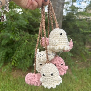 May include: A set of crocheted ghost and pumpkin ornaments hanging from brown strings. The ghosts are white with black eyes and the pumpkins are pink.