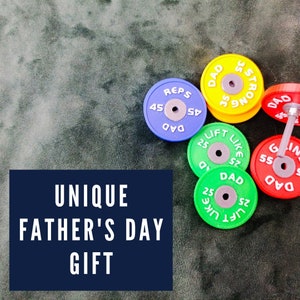 Könnte beinhalten: Ein Satz bunter Gewichtsplatten mit den Worten "Dad", "Reps", "Lift Like" und "Strong" darauf gedruckt. Die Platten sind ein einzigartiges Geschenk zum Vatertag.