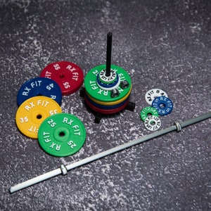 May include: A set of colorful weight plates with the text "RX FIT" and numbers printed on them. The plates are stacked on a black stand and a silver barbell is lying on a gray surface.