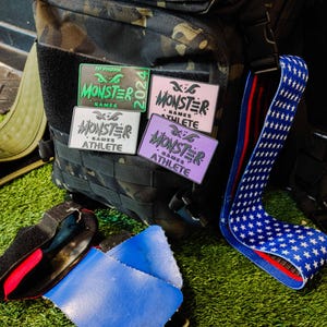 May include: Four different patches with the text "Monster Games Athlete" in various colors, including green, pink, white, and purple. The patches are attached to a black camouflage backpack with a red, white, and blue star patterned strap.