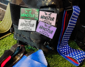 Può includere: Quattro patch diversi con il testo "Monster Games Athlete" in vari colori, tra cui verde, rosa, bianco e viola. I patch sono attaccati a uno zaino mimetico nero con una cinghia a stelle rosse, bianche e blu.