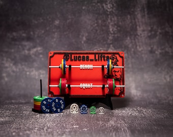 Puede incluir: Un rack de levantamiento de pesas rojo y negro con el texto "@Lucas_Lifts" y las palabras "Bench" y "Squat" impresas en él. El rack tiene dos barras de metal con placas de peso de colores. También hay varias placas de peso individuales en la mesa debajo del rack.