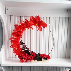 Pode incluir: Uma coroa de penas vermelhas com pequenas rosas vermelhas e folhagem em uma base de madeira. A coroa é decorada com pequenas luzes brancas e é exibida em uma prateleira branca.