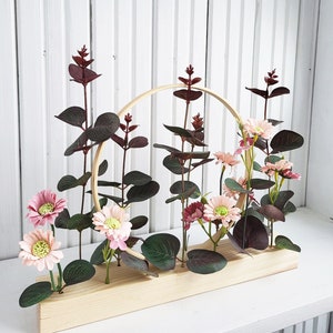 Op de afbeelding: Een houten basis met een ronde ring, versierd met roze en bordeauxrode kunstbloemen en groen.