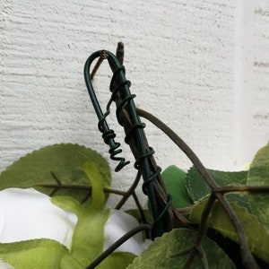 May include: Close-up of a green wire wrapped around a brown twig, with green leaves and a white fabric flower in the background.