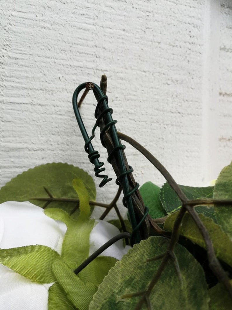 May include: Close-up of a green wire wrapped around a brown twig, with green leaves and a white fabric flower in the background.
