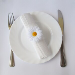 May include: A white plate with a white napkin tied with a white daisy with a yellow centre. A fork and knife are on either side of the plate.