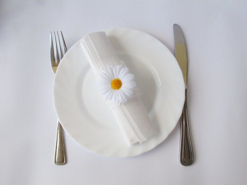 May include: A white plate with a white napkin tied with a white daisy with a yellow centre. A fork and knife are on either side of the plate.