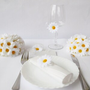 May include: White table setting with a white napkin, a white plate, a fork, a knife, and a glass with a white daisy. There are three white daisy arrangements on the table.